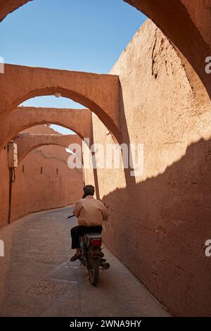 Un uomo in moto corre in una strada stretta con archi sopra nello storico quartiere Fahadan di Yazd, Iran. Foto Stock