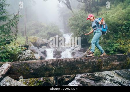 Giovane donna con zaino e bastoni da trekking che attraversa un ponte di legno vicino alla cascata del fiume Power Mountain durante il trekking al Parco Nazionale Makalu Barun nel NEPA Foto Stock