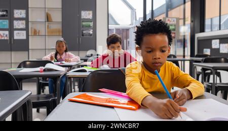 Il ragazzo birazziale in giallo si concentra sulla scrittura alla sua scrivania in una classe scolastica Foto Stock