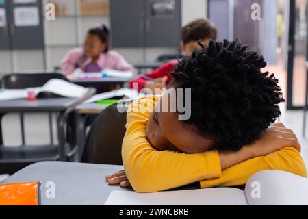 Un ragazzo birazziale in una camicia gialla poggia la testa sulle braccia alla scrivania di una scuola Foto Stock