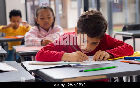Il ragazzo birazziale in rosso si concentra intensamente sul suo disegno in un ambiente scolastico Foto Stock