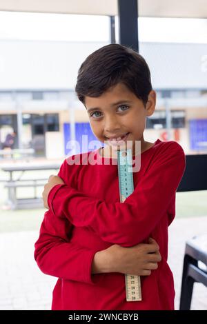 Un ragazzo birazziale con un sorriso brillante regge un sovrano, indossando una camicia rossa a scuola Foto Stock