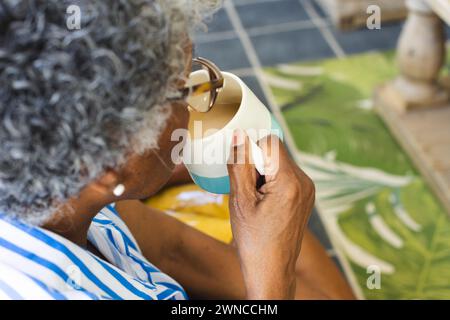 Una donna afroamericana anziana si gode una tazza di tè a casa, i suoi capelli grigi visibili da dietro Foto Stock