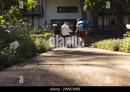 Donna birazziale anziana e uomo birazziale camminano verso una casa, tirando una valigia, con spazio per le copie Foto Stock