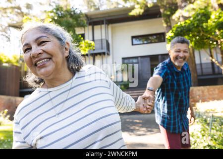 La coppia birazziale senior gode di un momento di gioco all'aperto, tenendosi per mano con espressioni gioiose Foto Stock