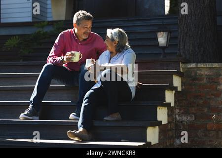 Una coppia birazziale senior gode di una giornata di sole sui gradini della veranda, condividendo un momento con un caffè Foto Stock