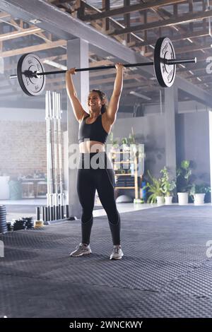 Una giovane donna birazziale in forma solleva un barbell in alto in un ambiente di palestra Foto Stock