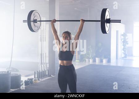 Una giovane donna birazziale dalla forma forte solleva un pesante campanile sopra la testa in una palestra Foto Stock