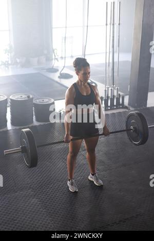 Una giovane donna **in forma birazziale** solleva un barbell in palestra, concentrandosi sul suo allenamento di forza Foto Stock