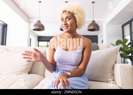 Giovane donna birazziale con capelli biondi ricci seduta sul divano, gestendo durante una videochiamata Foto Stock