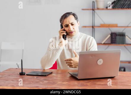 Giovane grafico uomo adulto che parla su un telefono cellulare in ufficio e lavora su un notebook e un tablet grafico. Foto Stock