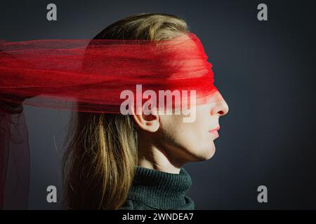 Visualizzazione di un problema psicologico. Ritratto di profilo di una giovane donna adulta con lunghi capelli biondi e una benda di tessuto rosso trasparente Foto Stock