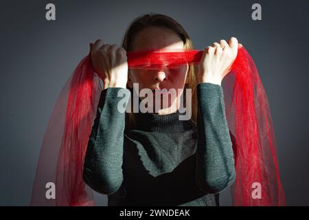 Visualizzazione di un problema psicologico. Una giovane donna adulta tiene una benda fatta di tessuto rosso trasparente sugli occhi. Foto Stock