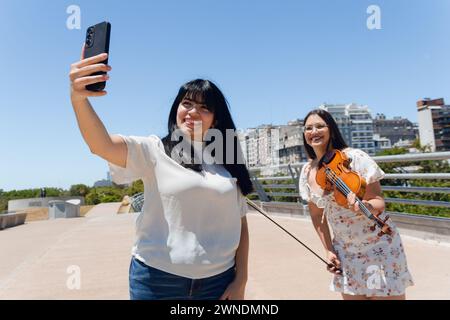 Giovane donna latina venezuelana all'aperto felice di scattare foto con il suo telefono con una violinista di strada con il suo violino, Foto Stock