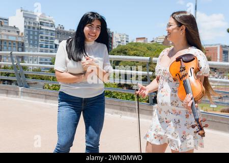 giovane donna turistica latino venezuelana che viaggia per buenos aires, felice di parlare per strada con la violinista busker donna a mezzogiorno, sorridere e chiacchierare Foto Stock