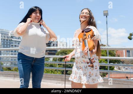 giovane donna turistica latino venezuelana che viaggia per buenos aires, felice di parlare per strada con la violinista busker donna a mezzogiorno, sorridere e chiacchierare Foto Stock