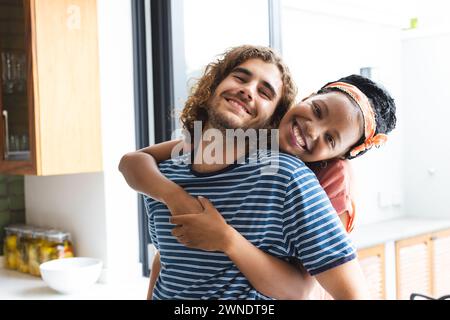 Coppia diversificata giovane donna afroamericana abbraccia un giovane uomo caucasico da dietro, entrambi sorridenti Foto Stock