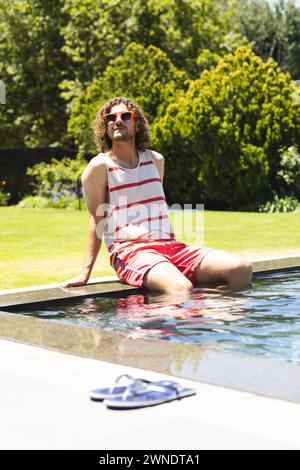 Giovane uomo caucasico con i capelli ricci siede vicino a una piscina, indossando occhiali da sole e abbigliamento estivo Foto Stock