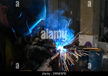 Saldatrice al lavoro. Scintille di saldatura. Lavoratore maschile che indossa casco e guanti. Saldatura del metallo in fabbrica. Primo piano del lavoratore industriale nel cantiere. Foto Stock