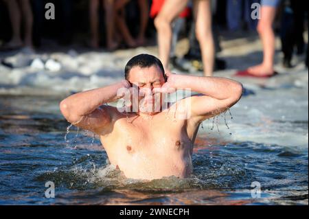 Battezzare. Giovane che stringe il naso prima di tuffarsi in un pozzo d'acqua fredda durante la festa Epifania sul fiume, le gambe della gente sullo sfondo. Januar Foto Stock