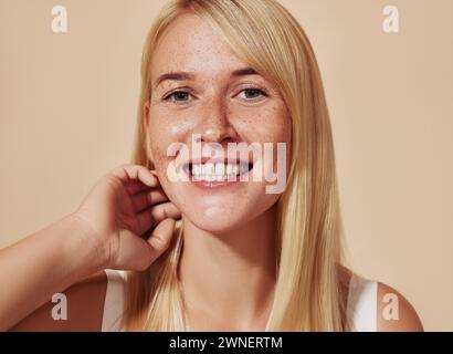 Giovane donna felice con capelli biondi che guarda la macchina fotografica in uno studio. Bella donna sorridente con lentiggini che si regolano i capelli su uno sfondo beige. Foto Stock