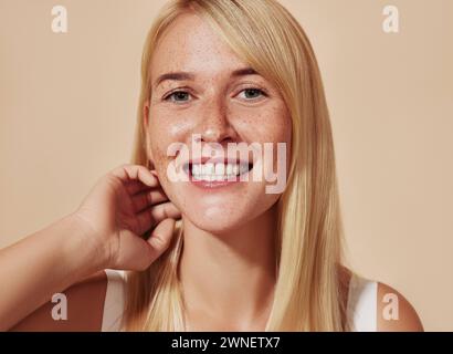 Giovane donna felice con capelli biondi che guarda la macchina fotografica in uno studio. Bella donna sorridente con lentiggini che si regolano i capelli su uno sfondo beige. Tu Foto Stock