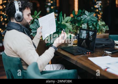 Uomo d'affari concentrato con cuffie che recensisce documenti in un ambiente accogliente. Foto Stock