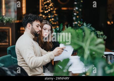 Giovani e donne concentrati collaborano alla creazione di documenti in un bar festoso, circondato da decorazioni e luci natalizie. Foto Stock