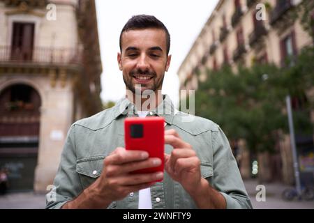 Giovane uomo attraente caucasico sta cercando felice cellulare per strada nella città di Barcellona. Foto Stock