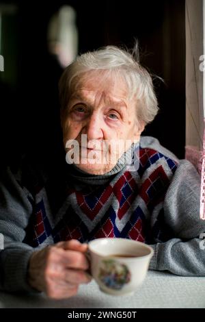 Una donna anziana siede a un tavolo con una tazza in mano. Foto Stock