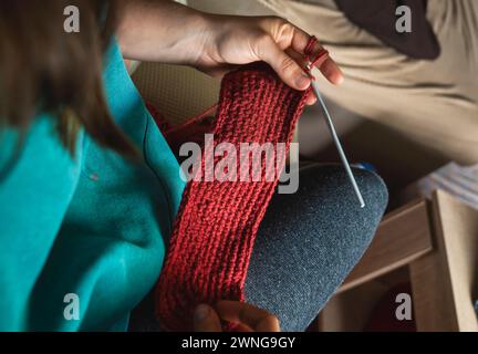 Giovane donna che lavora a maglia una sciarpa circolare con filato rosso. Seduta sul divano del soggiorno, primo piano sulle spalle. Foto Stock