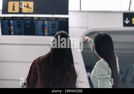 due ragazze multiculturali da dietro nel terminal dell'aeroporto puntano le dita sul display delle informazioni mentre cercano il volo Foto Stock