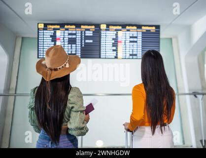 Da dietro due ragazze latine che viaggiano all'aeroporto con i bagagli in cerca del volo sulla bacheca informativa, una di loro con un cappello Foto Stock