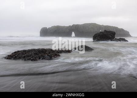 Un litorale roccioso con una grande formazione rocciosa in primo piano. L'acqua è instabile e il cielo è coperto, creando un'atmosfera allegra e drammatica Foto Stock