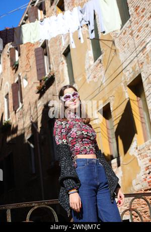 ragazza con maschera veneziana che indossa pantaloni blu jeans in posa vicino a case con abiti appesi Foto Stock