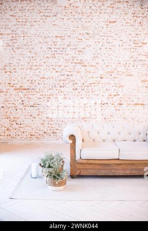 Interni di casa con un divano trapuntato bianco con base in legno davanti a una parete vuota in mattoni, con tende bianche e vasi di fiori su un tappeto grigio Foto Stock