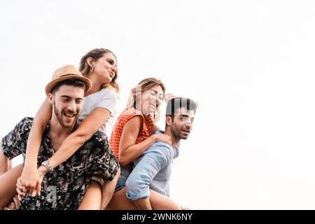 Due coppie gioiose che si impegnano in giri in piggyback, condividono risate contro un cielo limpido, incarnano l'amicizia e il divertimento: Amici felici che legano il bac bianco Foto Stock