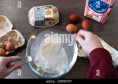 Le mani di un uomo tengono un setaccio e setacciano la farina, con ingredienti per cuocere una torta su un piano di lavoro della cucina: Burro, uova e farina. REGNO UNITO Foto Stock