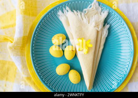 Questo vivace tavolo pasquale presenta un piatto blu adornato con uova gialle e un delicato fiore. La tovaglia a scacchi aggiunge un tocco natalizio Foto Stock