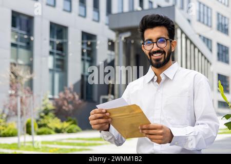 Un uomo d'affari allegro e barbuto che indossa gli occhiali legge una lettera all'aperto. Sorride in un elegante abbigliamento casual, con moderni edifici per uffici che fanno da sfondo a una scena di successo professionale e corrispondenza positiva. Foto Stock