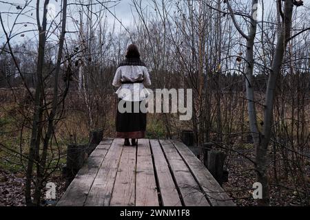 Una giovane donna in abiti antichi si trova su una piattaforma vicino a un vecchio stagno. La ragazza dorme vicino a una palude sporca con cespugli, copia spazio Foto Stock