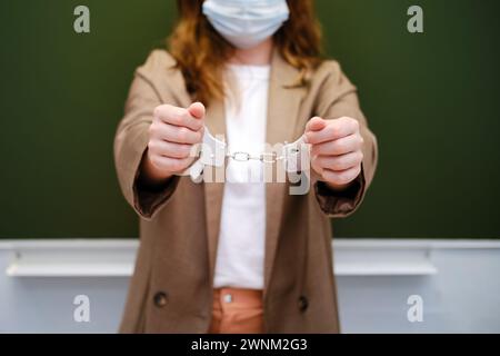 Insegnante di scuola in maschera di faccia incatenata da manette si trova al lavagna, concetto di problema epidemico Foto Stock