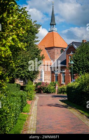 Un sentiero acciottolato conduce a una piccola chiesa in mattoni in una giornata di sole con un confine verde con siepi, Rysum, Krumhoern, Frisia orientale, bassa Sassonia Foto Stock