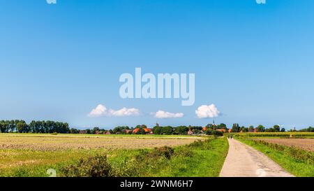 Una strada di campagna conduce attraverso tranquilli campi rurali con un villaggio in lontananza sotto un cielo azzurro, Rysum, Krumhoern, Frisia orientale, bassa Sassonia Foto Stock