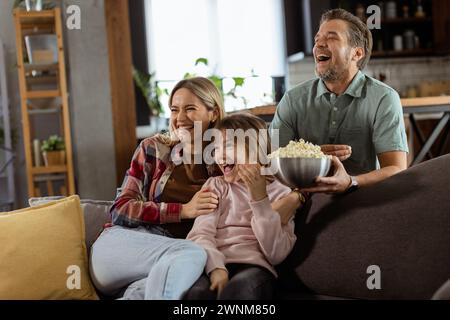 Una famiglia di tre persone si trova comodamente adagiata su un divano, con i volti che riflettono l'emozione e l'attenzione mentre condividono una ciotola di popcorn durante una sosp Foto Stock