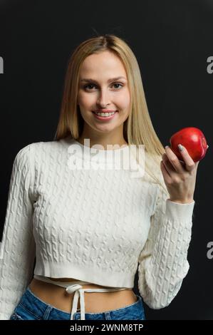 Bella giovane donna con una mela rossa su sfondo nero. Ragazza bionda con bretelle in un maglione bianco. Foto Stock