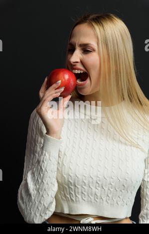 Bella giovane donna con una mela rossa su sfondo nero. Ragazza bionda con bretelle in un maglione bianco. La ragazza morde una mela. Foto Stock