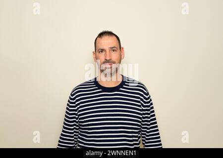 Uomo ispanico con barba che indossa un maglione a righe che sorride sullo sfondo beige dello studio. Foto Stock