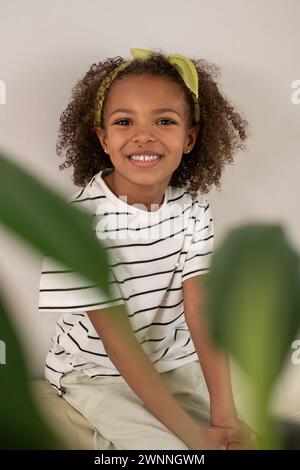 Una allegra ragazza mista con una camicia a righe e capelli ricci legati con un cerchietto visto attraverso foglie verdi sfocate, sorriso e felicità, National Sib Foto Stock