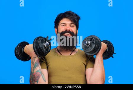 Uomo barbuto sorridente con manubri in mano. Atleta forte che si allena nella palestra fitness. Uomo muscoloso atletico che solleva manubri nel club sportivo Foto Stock